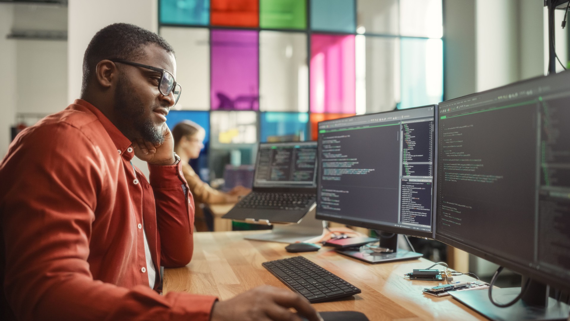 Black Man Writing Lines of Code On Desktop PC With Two Monitors and a Laptop in Stylish Office. Professional Male Developer Programming Artificial Intelligence Software for Start-Up Company.